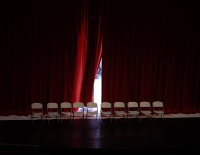 A red stage curtain is parted slightly showing light behind it. A row of white metal chairs is in front of the curtain on the black stage. 
