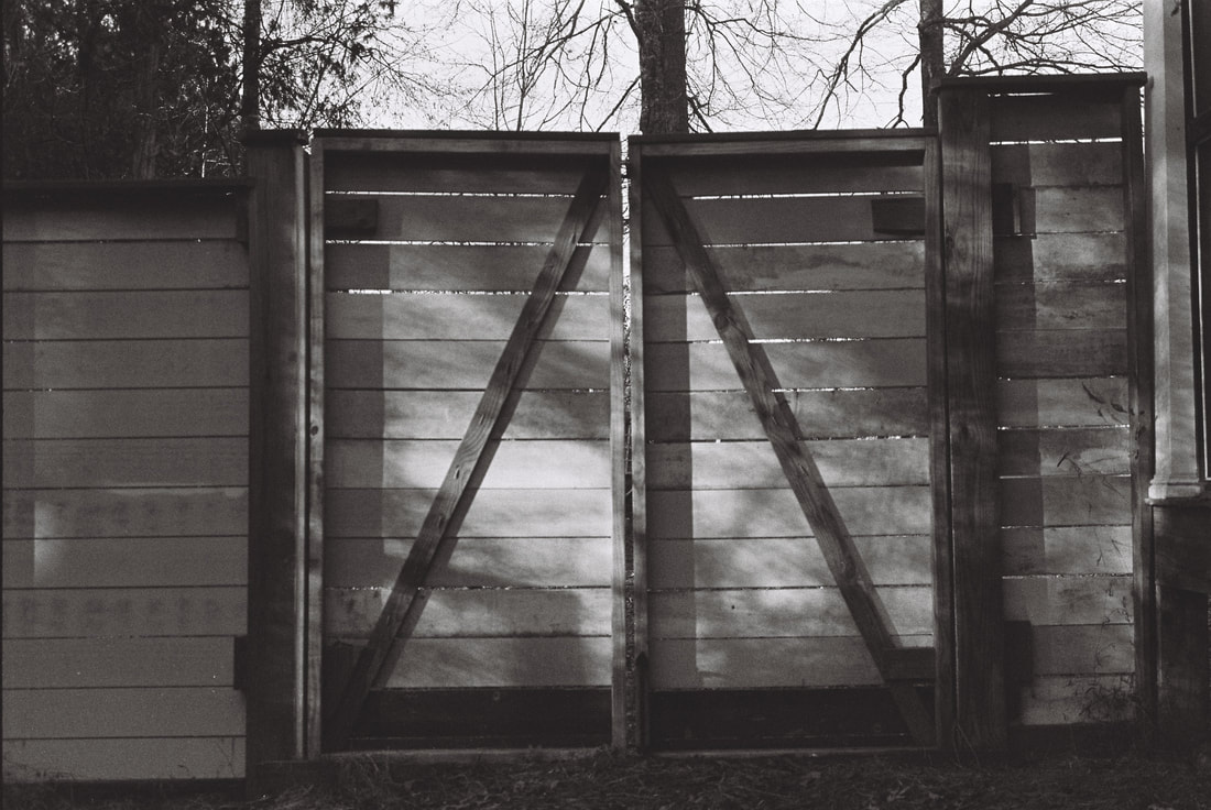 A wooden gate constructed with horizontal slats is photographed in black and white with shadows being cast upon it from late afternoon sun.  Trees and sky are on the other side.